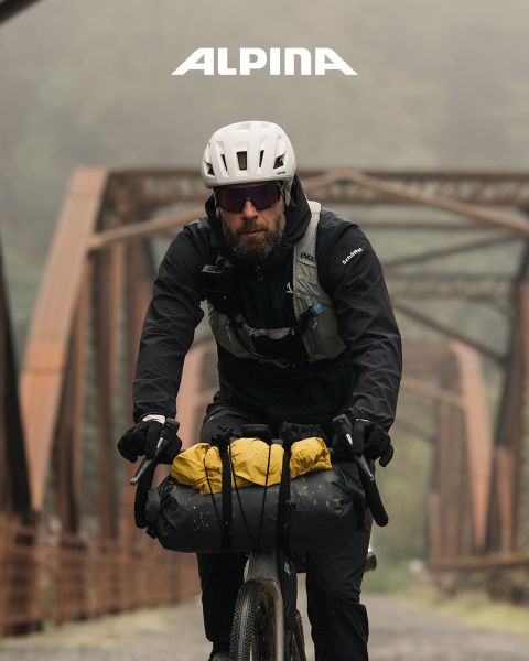 Un ciclista con casco y camisa blanca está de pie con su bicicleta mirando a la distancia al atardecer. En el fondo se ven árboles, colinas y una línea eléctrica. Encima se ve la palabra "ALPINA".