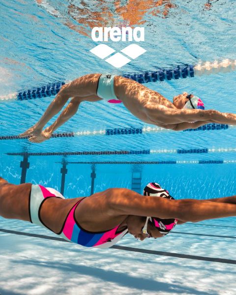 Un nadador con gorro de natación negro y gafas de natación reflectantes se desliza bajo el agua en una piscina marcada con separadores de carriles. El traje de baño azul tiene adornos naranjas. El agua está clara y, por encima, se ve el logotipo de Arena.
