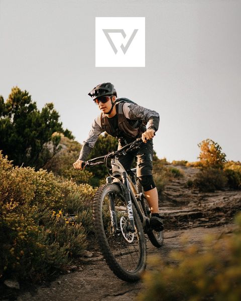 Dos ciclistas montan en bicicleta de montaña por un sendero accidentado en una región montañosa con picos nevados al fondo. Llevan cascos y equipo para actividades al aire libre. En la parte superior de la imagen se ve un logotipo.