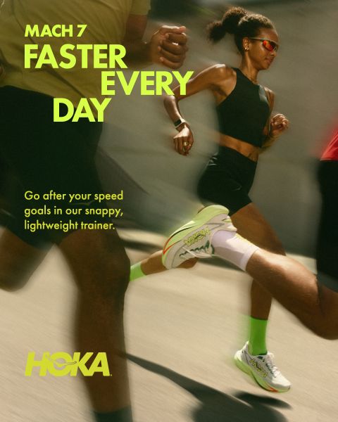 Dos corredores, un hombre y una mujer, corren por un sendero rocoso de montaña, rodeados de exuberante vegetación y altos árboles. Ambos llevan ropa de correr y mochilas de hidratación. En la parte superior se muestra claramente la palabra "HOKA". En el fondo se alzan montañas nevadas.