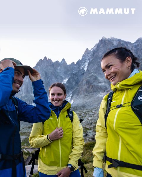 Dos excursionistas con equipo de exterior atraviesan un terreno rocoso bajo un cielo nublado, con una gran montaña al fondo. Ambos visten ropa de Mammut, y el logotipo de la marca se ve en la parte superior de la imagen.