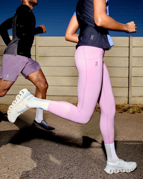 Una persona corre al aire libre. Lleva una chaqueta cortavientos beige-gris, pantalones cortos gris claro y zapatillas blancas para correr. El fondo es un cielo despejado y una superficie plana. El logo "On" es visible en la esquina superior derecha.
