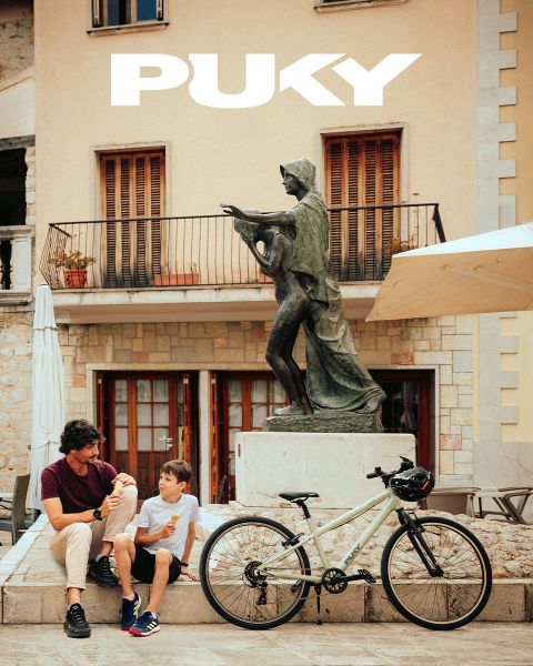 Una joven con casco azul y camiseta a rayas monta en bicicleta celeste en un día soleado. Sonriendo, pasa junto a coloridos grafitis en un entorno urbano. El logo de PUKY y el eslogan "Preparados para la vida" se ven en la parte superior izquierda.