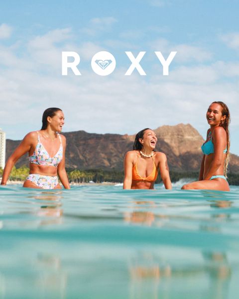 Tres mujeres nadan bajo el agua en trajes de baño en agua clara y azul. Una lleva una camisa de manga larga, las otras bikinis. La imagen está superpuesta con el logo blanco de "Roxy".