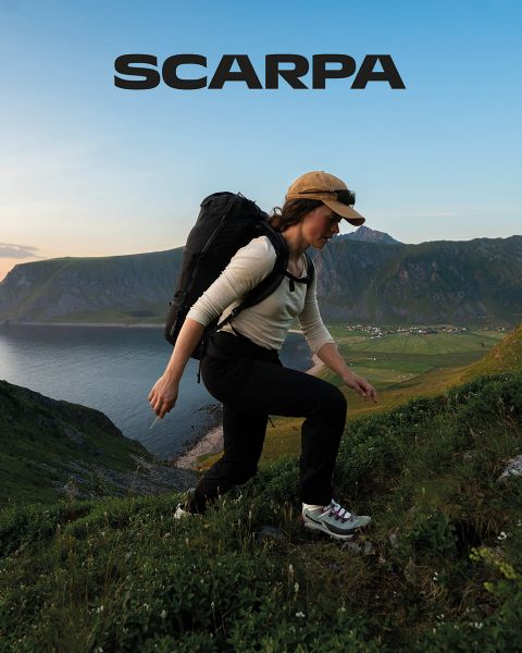 Una mujer y un hombre están sentados al aire libre sobre grandes rocas. Llevan ropa casual y gafas de sol. Están rodeados de vegetación y rocas, el cielo está ligeramente nublado. En la parte superior de la imagen se puede ver la palabra "SCARPA".