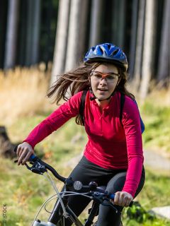 Una mujer con una camisa roja de manga larga, un casco y gafas anda en bicicleta en un área cubierta de hierba y arbolada en un día soleado.