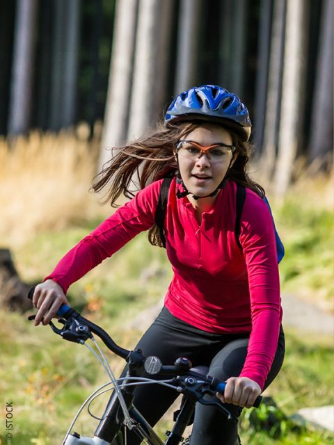 Una mujer con una camisa roja de manga larga, un casco y gafas anda en bicicleta en un área cubierta de hierba y arbolada en un día soleado.