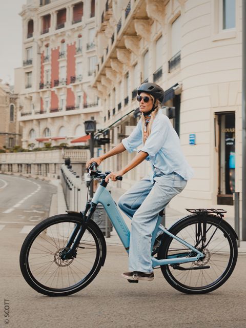 Una persona con casco y gafas de sol monta en una bicicleta azul claro en una calle urbana bordeada de edificios históricos.