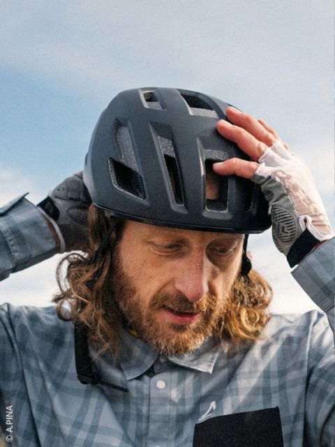 Un hombre con pelo largo y barba se ajusta su casco de bicicleta negro, lleva una camisa a cuadros y guantes sin dedos, en el fondo se ve el cielo.