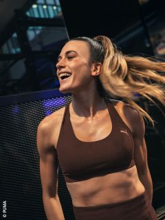 Una mujer con un sujetador deportivo marrón sonríe alegremente mientras hace ejercicio y su cabello se agita a un lado en una coleta alta.