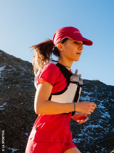 Una mujer en ropa de correr roja trota con un chaleco de hidratación bajo un cielo azul en una ruta de montaña.