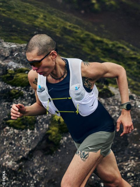 Una persona con tatuajes camina cuesta arriba por un camino pedregoso y cubierto de musgo, lleva gafas de sol, un chaleco para beber y un reloj inteligente.