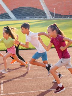 Cuatro niños corren en una pista en un estadio, sonríen y disfrutan de la carrera en un día soleado.