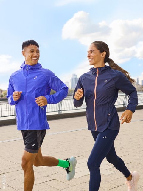 Dos personas en ropa deportiva trotan al aire libre por un camino pavimentado junto a un río y se sonríen, con el horizonte de la ciudad y el cielo azul al fondo.