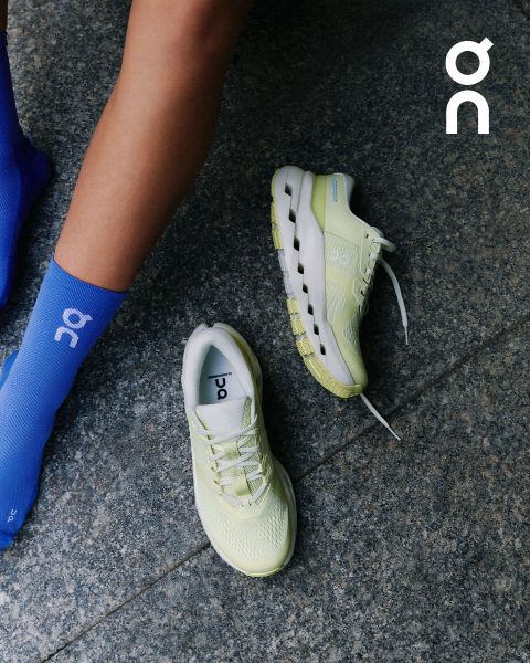 Una persona con calcetines azul claro junto a un par de zapatillas de correr ON de color amarillo claro sobre un suelo de piedra gris, con el logotipo de ON en la esquina superior derecha.