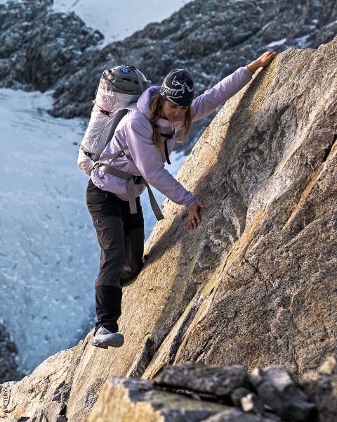Una mujer con mochila escala una ladera empinada y rocosa de montaña. Lleva equipo de exterior y un sombrero, mientras que en el fondo se ven picos nevados.