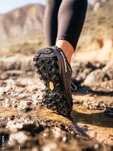 Primer plano de una persona con zapatillas de trail running negras corriendo sobre un terreno rocoso y húmedo al aire libre, con montañas al fondo desenfocado.