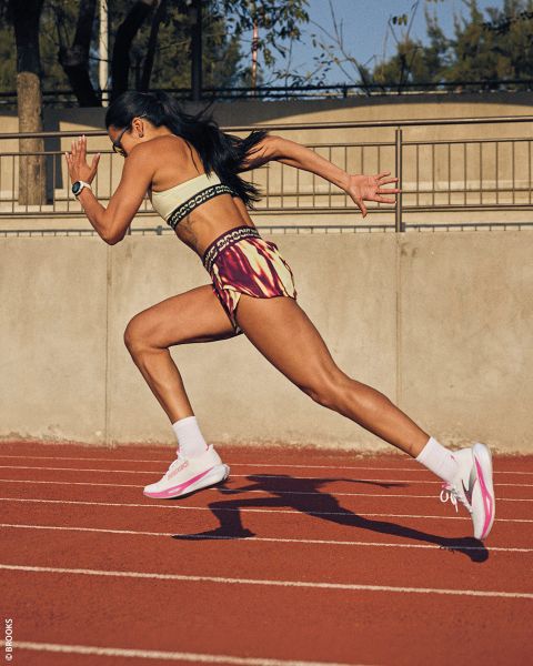 Una mujer en ropa deportiva corre en una pista roja al aire libre, con los brazos bombeando y el cuerpo inclinado hacia adelante en medio del paso.