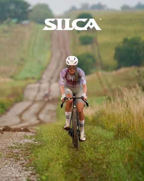 Ciclista subiendo una carretera de grava a través de campos cubiertos de hierba. Lleva casco y ropa de ciclista, con el logo de "SILCA" en la parte superior de la imagen.