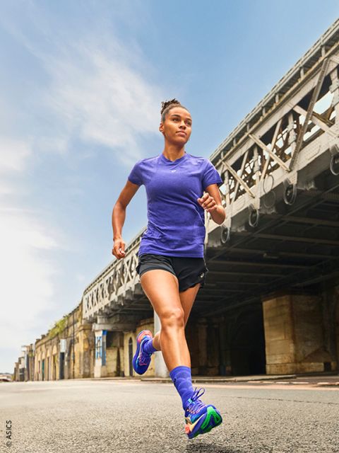 Una mujer en ropa deportiva corre al aire libre en un día claro en una calle de la ciudad debajo de un gran puente de metal.