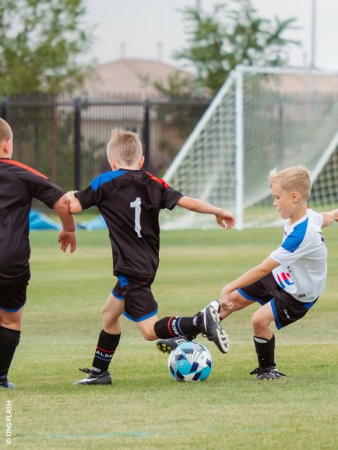 Tres niños juegan al fútbol en un prado; uno de blanco patea el balón, mientras que dos de negro intentan bloquearlo cerca de la portería.