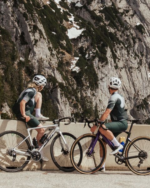 Dos ciclistas con ropa a juego se detienen en una barrera de carretera de montaña, con acantilados rocosos y manchas de nieve al fondo.