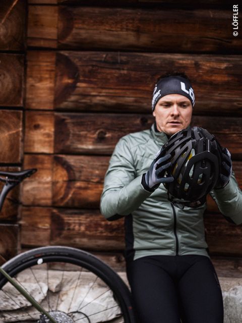 Un ciclista con una chaqueta verde y un gorro negro inspecciona un casco mientras se apoya en una pared de madera junto a una bicicleta.