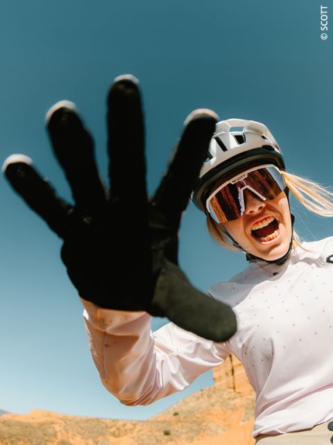 Un ciclista con casco y gafas de sol sonríe enérgicamente y extiende una mano enguantada hacia la cámara frente a un cielo azul despejado.