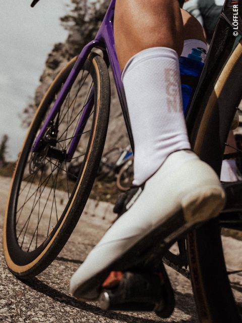 Primer plano de la pierna y el zapato blanco de un ciclista en una bicicleta morada, con una carretera borrosa y un paisaje rocoso al fondo.