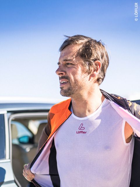 Un hombre al aire libre, que lleva una camiseta de rejilla blanca y una chaqueta naranja, está de pie junto a un coche bajo un cielo azul despejado.