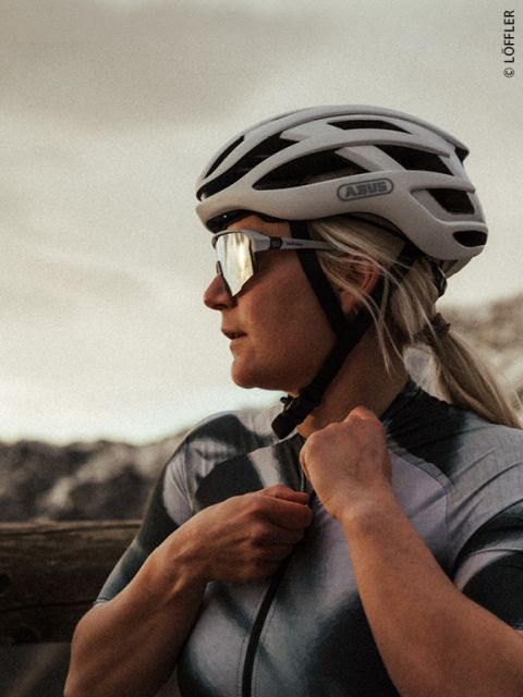 Una ciclista con casco y gafas de sol se ajusta el maillot al aire libre, con montañas al fondo al atardecer.