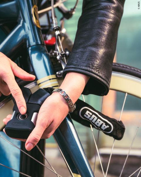 Una persona con una chaqueta de cuero cierra una bicicleta azul con un candado ABUS, concentrándose en asegurar el cuadro y la rueda.
