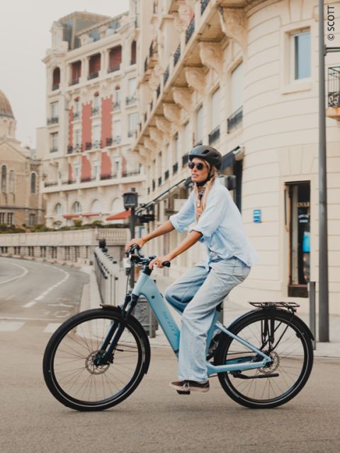 Una mujer con casco y gafas de sol monta en una bicicleta azul por una calle bordeada de edificios elegantes.