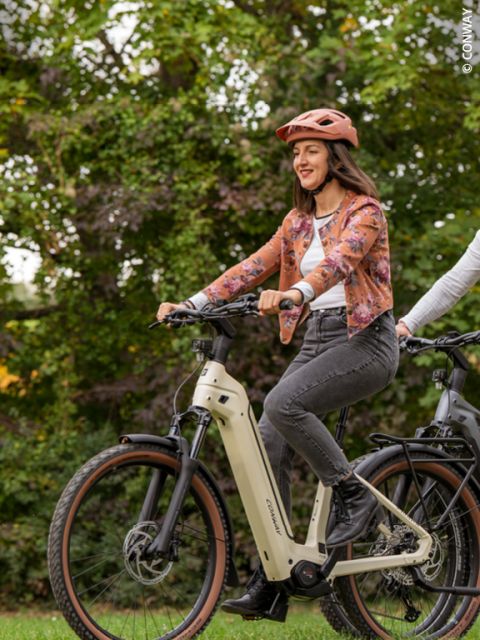 Una mujer que lleva un casco y una chaqueta floreada sonríe mientras monta en una bicicleta eléctrica al aire libre, con zonas verdes al fondo.