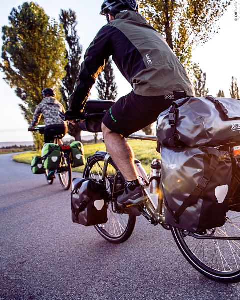 Dos ciclistas con alforjas viajan por una carretera adoquinada rodeada de árboles e iluminada por la cálida luz del sol.