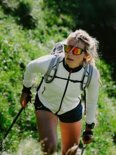Una mujer camina cuesta arriba por un sendero verde, lleva gafas de sol, una chaqueta blanca, una mochila y usa bastones de trekking.