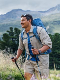 Un hombre sonriente con pelo largo y barba, vestido con una camisa azul y una mochila, de pie al aire libre frente a un fondo natural borroso.