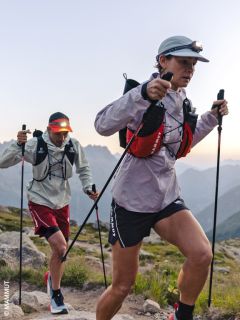 Dos excursionistas con linternas frontales y bastones de trekking ascienden por un sendero rocoso de montaña al amanecer, con ropa deportiva y mochilas, con las montañas al fondo.