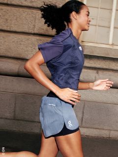 Mujer trota al aire libre en ropa deportiva, sonríe y pasa corriendo junto a un edificio de piedra con ventanas.