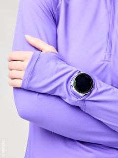 Una persona con una camisa de manga larga morada cruza los brazos y lleva un reloj inteligente negro redondo sobre la manga.