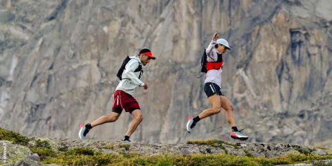 Dos personas haciendo trail running en terreno rocoso con una gran pared de roca al fondo, ambos con mochilas, gorras y ropa deportiva.