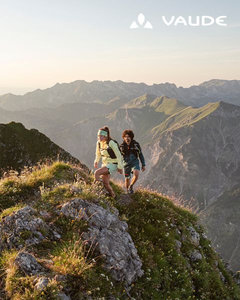 Dos excursionistas con mochilas trepan por una cresta montañosa cubierta de hierba al amanecer, con montañas escarpadas al fondo. El logotipo de Vaude aparece en la esquina.