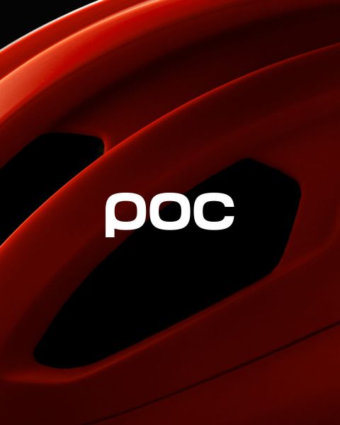 Una persona con un abrigo de invierno naranja y un casco está de pie en la cima de una montaña nevada, mirando las lejanas montañas cubiertas de nieve. El logo de POC se puede ver en el casco y en la esquina superior derecha de la imagen.