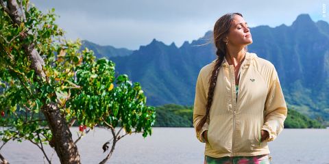 Una mujer con una chaqueta beige está de pie junto a un lago con montañas al fondo, con los ojos cerrados y la cara hacia el sol, junto a un árbol frondoso.