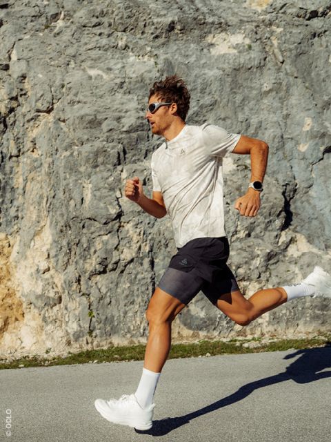 Un hombre con gafas de sol y ropa deportiva camina bajo la brillante luz del sol por una calle que pasa junto a una pared rocosa.