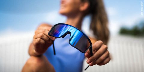 Una mujer con un top azul sostiene unas gafas de sol deportivas con espejo hacia la cámara, el fondo exterior está desenfocado.