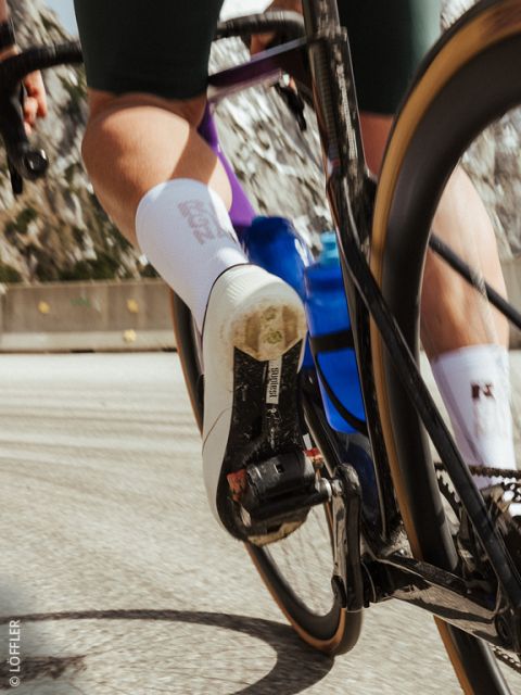 Primer plano del pie de un ciclista en un pedal, con zapatilla de ciclismo blanca, botella de agua azul y movimiento borroso en una carretera.