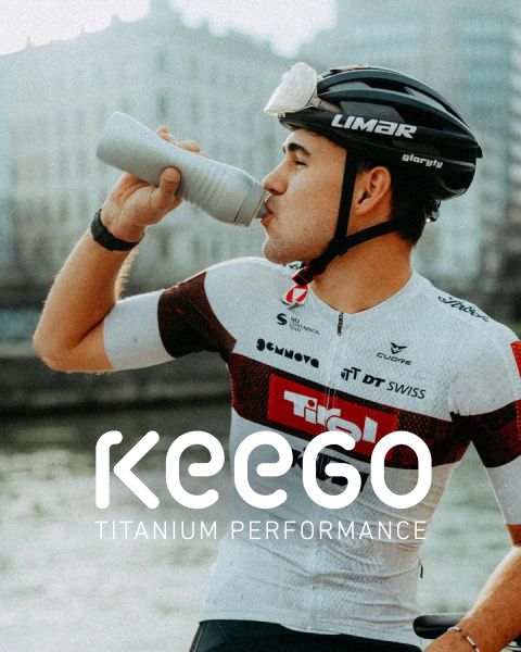 Un ciclista con un maillot blanco y rojo bebe de una botella y lleva un casco en el que se muestra el texto "KEEGO Titanium Performance".