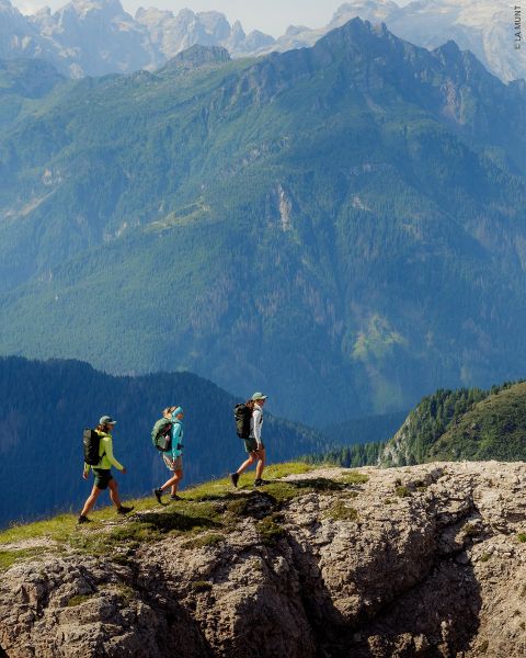 Tres excursionistas con mochilas caminan a lo largo de una cresta rocosa con altas montañas boscosas al fondo bajo un cielo despejado.