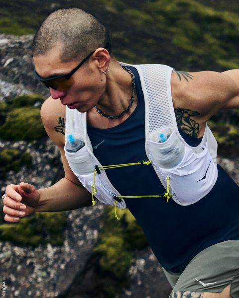 Una mujer en ropa deportiva y con gorra lleva un chaleco para correr con botellas de agua y está de lado frente a un fondo blanco sencillo.